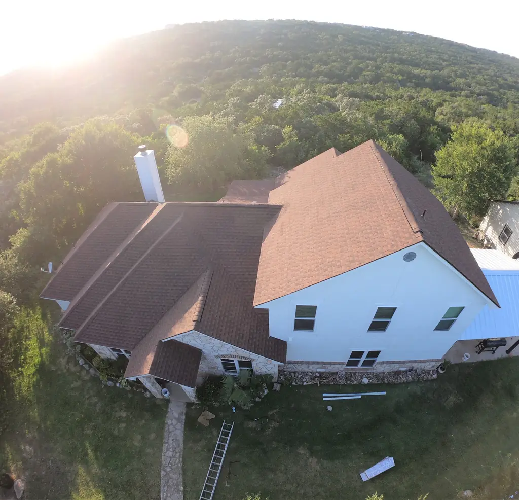 Golden hour aerial of a Hill Country home near San Antonio with a completed brown shingle roof and a ladder still against the eave
